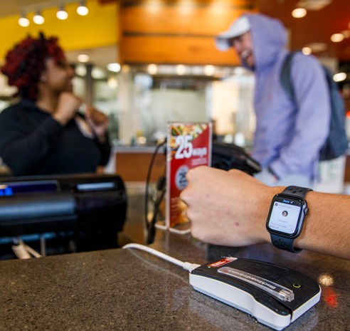 Bama Dining cashier laughs with student in the background while another student taps their ACT Card for a meal swipe